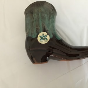May include: A ceramic cowboy boot figurine, primarily brown with a glossy finish. The top of the boot is a mottled green. A sticker with the text "1867 1967" is affixed to the boot. The boot is a decorative item.