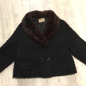May include: A vintage black jacket with a dark brown fur collar. The jacket has a textured fabric, two front pockets, and two black buttons. The label inside the jacket is visible.