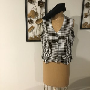 May include: A black and white gingham sleeveless vest with a V-neckline and button closure. The vest has two pockets on the front and is displayed on a mannequin.