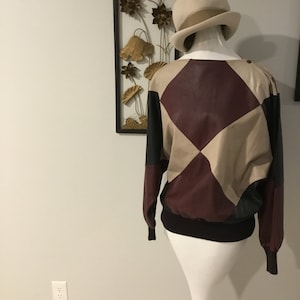 May include: A brown, black, and beige leather jacket with a diamond pattern. The jacket has a ribbed knit collar, cuffs, and hem. The jacket is on a white mannequin.