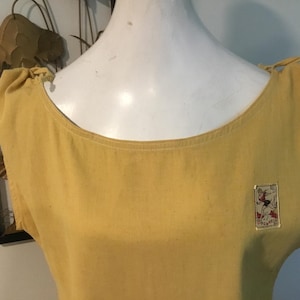May include: Mustard yellow sleeveless top with a scoop neckline and shoulder ties. A small rectangular patch with a graphic illustration is affixed to the front. The top is displayed on a white mannequin.