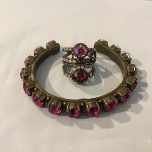 May include: A gold-toned metal cuff bracelet and three rings. The cuff bracelet is adorned with pink gemstones. The rings feature pink and clear gemstones. Jewellery.
