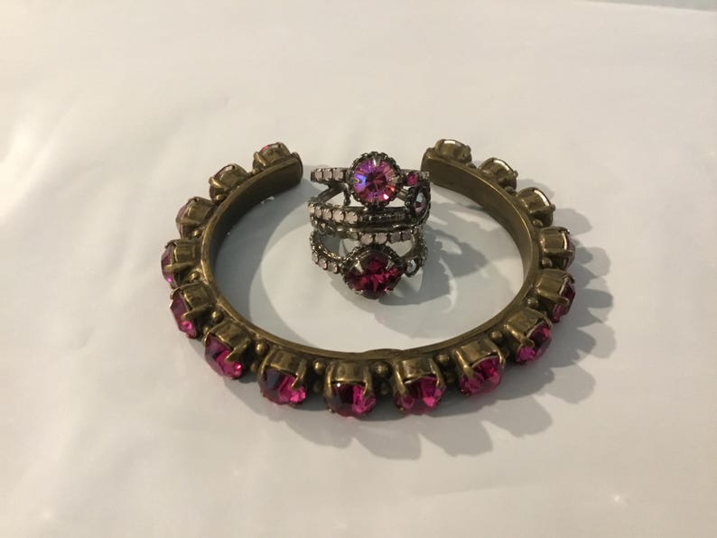 May include: A gold-toned metal cuff bracelet and three rings. The cuff bracelet is adorned with pink gemstones. The rings feature pink and clear gemstones. Jewellery.