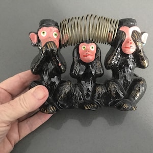 May include: Three black ceramic monkeys with pink faces, each covering a different part of their face. The monkey on the left is covering its mouth, the middle monkey is covering its eyes, and the monkey on the right is covering its ears. The monkeys are sitting on a spring.