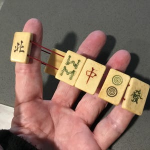 May include: A bracelet constructed from five rectangular off-white mahjong tiles strung together with thin red elastic cord. Each tile features distinct black, green, or red engraved Chinese characters and circular symbols on the front surface.