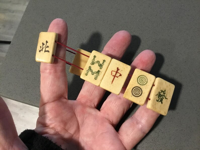 May include: A bracelet constructed from five rectangular off-white mahjong tiles strung together with thin red elastic cord. Each tile features distinct black, green, or red engraved Chinese characters and circular symbols on the front surface.