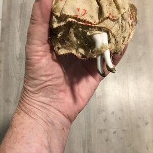 May include: A vintage doll with porcelain legs and feet, wearing a cream-colored dress with red accents. The doll's legs are white with gold-painted shoes. The doll is held in a hand.