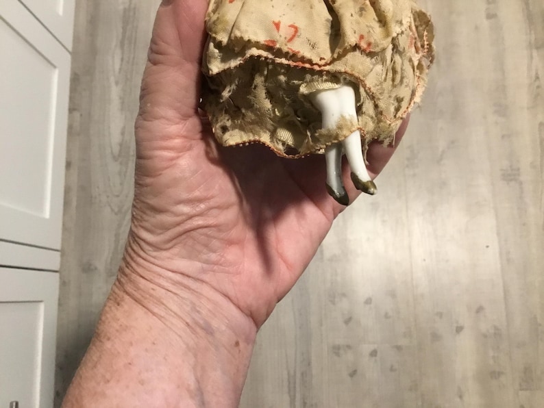 May include: A vintage doll with porcelain legs and feet, wearing a cream-colored dress with red accents. The doll's legs are white with gold-painted shoes. The doll is held in a hand.