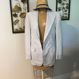 May include: A light blue blazer with white stripes, worn on a mannequin with a straw hat. The blazer has a single button closure and two pockets.