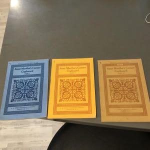 Peut inclure: Trois livrets vintage intitulés "Aunt Martha's Corner Cupboard" sont présentés. Les couvertures sont bleues, jaunes et beiges, chacune avec un motif floral décoratif et le texte "Part I", "Part II" et "Part III". Les livrets proviennent de F.A. Owen Publishing Company.