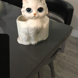 May include: A white ceramic cat planter with large blue eyes and a small orange nose. The cat is sitting upright with its paws together. The planter has a cylindrical base.