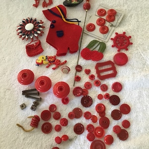 May include: A collection of red buttons, beads, and other craft supplies. There are a variety of shapes and sizes, including round, square, and star-shaped buttons. There are also some small red beads and a red felt doll.