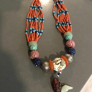 May include: A multi-strand necklace featuring orange, blue, and turquoise beads. The necklace includes carved stone beads in various colors and a central pendant with a white carved figure, orange accents, and a dangling charm with a red coral drop.