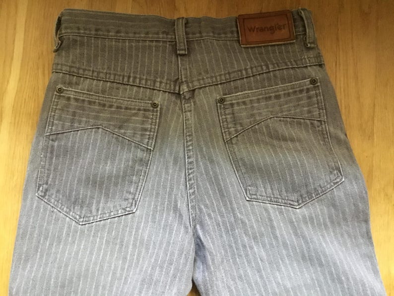 K&ouml;nnte beinhalten: Graue gestreifte Jeans mit einem braunen Leder-Patch, auf dem "Wrangler" auf der R&uuml;ckseite steht.