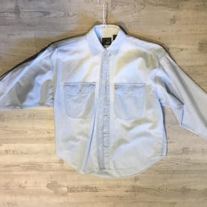 May include: Light blue denim shirt with long sleeves, two chest pockets, and button-down closure. The shirt is hanging on a white hanger against a light wood grain background. A classic, casual style.