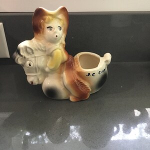 May include: A ceramic planter shaped like a cowboy riding a horse. The planter is painted in shades of brown, white, and yellow. The cowboy has a brown hat and a blue shirt. The horse has a brown mane and tail. The planter has a small opening on the top for planting. The planter has the text "S.C. Craft" on the side.