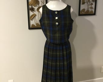 May include: A vintage plaid dress and hat set. The sleeveless dress features a green, blue, and black plaid pattern with a pleated skirt and three white buttons. A black hat with a white lace trim sits atop a mannequin head.