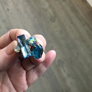 May include: A cluster ring featuring multiple blue gemstones. The ring has a rectangular, oval, and round-cut stones in varying shades of blue and iridescent hues. The ring is set on a silver-toned band.
