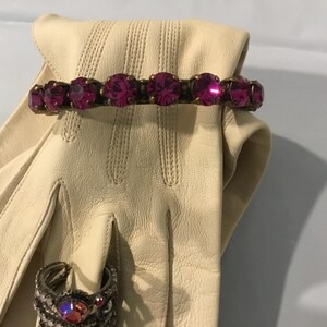 May include: A cream leather glove with a magenta rhinestone bracelet and a silver ring with a magenta stone. The bracelet has seven large, round magenta rhinestones set in a gold-toned metal.