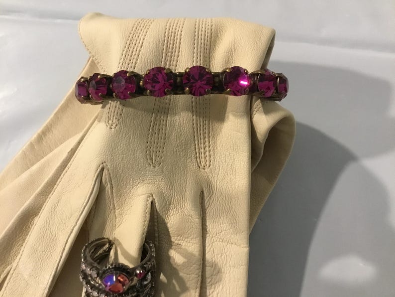 May include: A cream leather glove with a magenta rhinestone bracelet and a silver ring with a magenta stone. The bracelet has seven large, round magenta rhinestones set in a gold-toned metal.