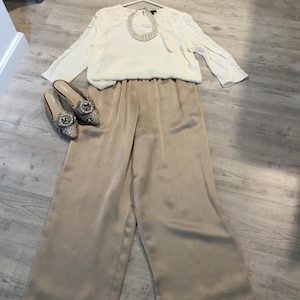 May include: A white long-sleeved blouse with lace shoulder detailing, a cream-colored pearl-style necklace, beige wide-leg trousers with an elastic waistband, and a pair of snakeskin-patterned loafers featuring large decorative crystal buckles.