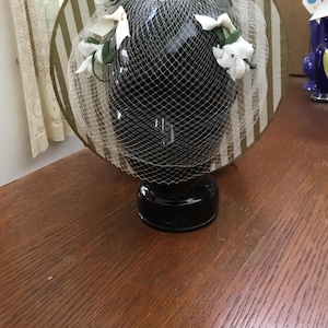 Peut inclure: Un chapeau vintage avec une base noire, orné de fleurs blanches et d'un voile délicat. Le chapeau présente un large bord avec des rayures verticales dorées et blanches. Le chapeau est présenté sur un support noir.