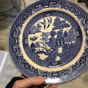 May include: A blue and white porcelain plate with a traditional willow pattern design. The plate features a central image of a pagoda, trees, and a river, surrounded by a decorative border.