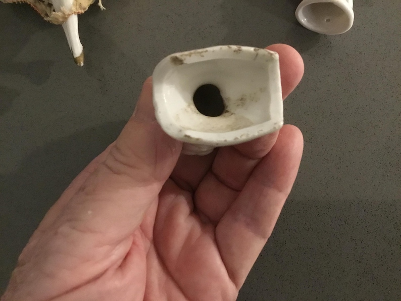 May include: A white porcelain doll head base with a dark hole. The base is held in a hand, with a doll leg and another doll part visible in the background. The porcelain has some discoloration.