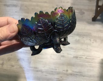 May include: A black iridescent glass bowl with a leaf design and three legs. The bowl has a shiny finish and is decorated with a swirling pattern of colors.