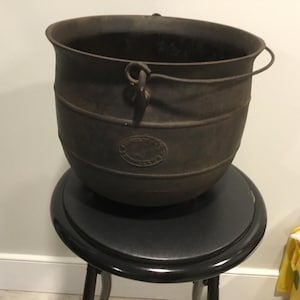 May include: A dark gray cast iron cauldron with a rounded body, a wide open top, and a thin metal wire handle attached to two side lugs. The exterior features a circular embossed logo on the front and horizontal ridges around the middle.
