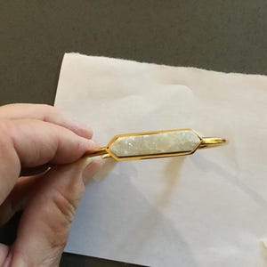 May include: Gold-toned bracelet with a rectangular, light-colored stone center. The stone has a textured, crystalline appearance, set within a gold frame. The bracelet is held against a white surface.