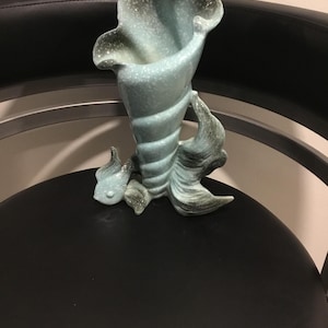 May include: A light blue ceramic vase in the shape of a conch shell. The vase has a textured surface with speckled detailing and a flared opening. The base features stylized fins, adding to the nautical theme. A decorative item for home decor.