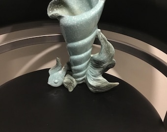 May include: A light blue ceramic vase in the shape of a conch shell. The vase has a textured surface with speckled detailing and a flared opening. The base features stylized fins, adding to the nautical theme. A decorative item for home decor.