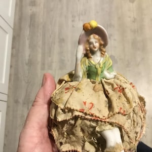 May include: A vintage porcelain doll with a bonnet and a floral patterned dress. The doll has a painted face and is wearing a green and yellow top. The doll's dress is beige with red floral accents.