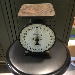 May include: A black vintage scale with a white face and black numbers. The scale has a round dial with markings for ounces and pounds. The scale is sitting on a black stool.