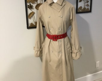 May include: A beige double-breasted trench coat with a wide collar, long sleeves with buckled cuffs, and a bright red patent leather belt with a rectangular buckle. A textured red beret sits atop the white mannequin wearing the coat.