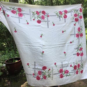 May include: A white tablecloth with a floral pattern of pink roses and green leaves. The tablecloth has a rectangular shape and is decorated with a border of roses and a grid design.