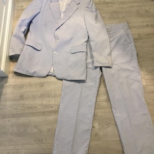 May include: A light blue and white striped suit, including a blazer and matching trousers, is laid out on a light wood-grain floor. The blazer has two front pockets and a single-button closure. The trousers are neatly folded.