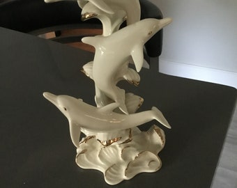 May include: A porcelain figurine of three white dolphins leaping out of the water. The dolphins are painted with gold accents and are arranged in a cascading formation. The base of the figurine is a white wave with gold accents.