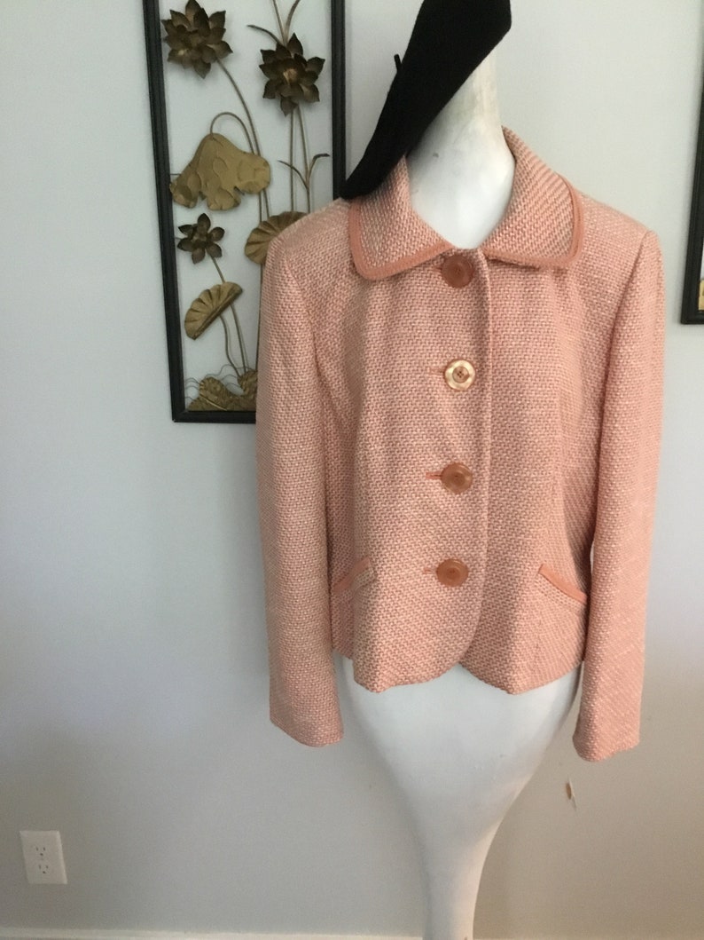 May include: A pink tweed blazer with a collared neckline and four button closures. The blazer has a fitted silhouette and a slightly cropped length.