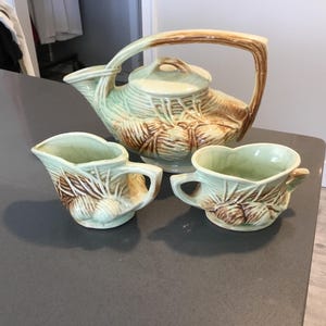 May include: A vintage ceramic tea set in shades of green and brown. The set includes a teapot with a large handle, a creamer, and a small cup. The pieces are decorated with a leaf and bulb design.