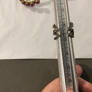 May include: A silver ring with an elaborate design, featuring multiple curved elements and small embellishments. The ring is being measured on a ring sizer, indicating a size 16. A second ring with pink gemstones is visible in the background.
