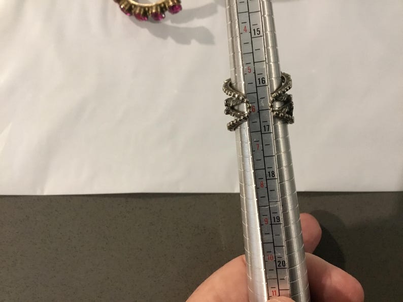 May include: A silver ring with an elaborate design, featuring multiple curved elements and small embellishments. The ring is being measured on a ring sizer, indicating a size 16. A second ring with pink gemstones is visible in the background.