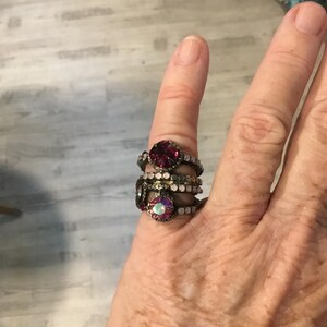 May include: A collection of decorative rings featuring ruby red and iridescent gemstones. The rings are stacked on a finger, highlighting the intricate design and vintage aesthetic of the jewellery.
