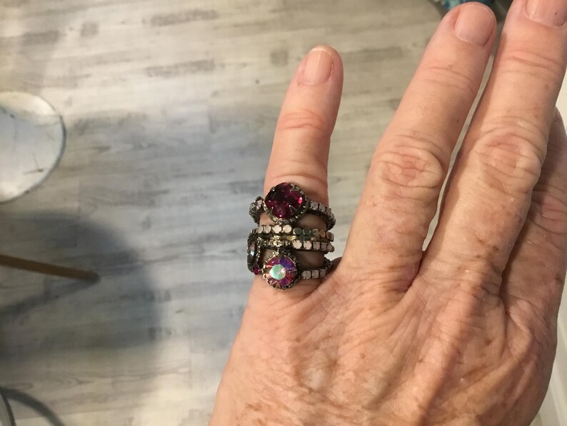 May include: A collection of decorative rings featuring ruby red and iridescent gemstones. The rings are stacked on a finger, highlighting the intricate design and vintage aesthetic of the jewellery.