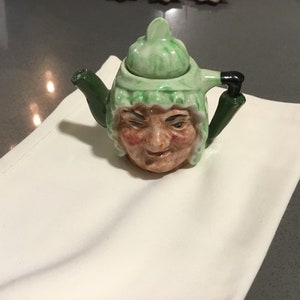 May include: A whimsical ceramic teapot with a face design. The teapot features a green hat and spout, with a face that has rosy cheeks and a slightly mischievous expression. The teapot is resting on a white cloth.