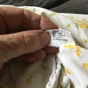Peut inclure: Un drap-housse blanc pour lit simple avec un motif floral jaune. L'&eacute;tiquette indique TWIN FITTED, 50% Polyester, 50% Cotton et JCPenney. Le drap est compos&eacute; d'un m&eacute;lange de polyester et de coton avec un imprim&eacute; de fleurs jaunes r&eacute;p&eacute;titives.