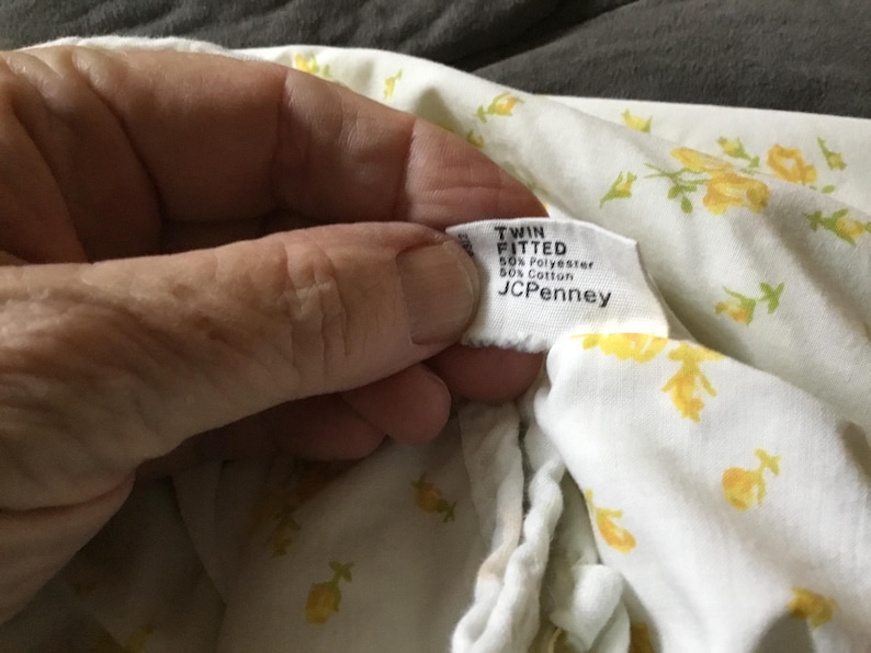 Peut inclure: Un drap-housse blanc pour lit simple avec un motif floral jaune. L'&eacute;tiquette indique TWIN FITTED, 50% Polyester, 50% Cotton et JCPenney. Le drap est compos&eacute; d'un m&eacute;lange de polyester et de coton avec un imprim&eacute; de fleurs jaunes r&eacute;p&eacute;titives.