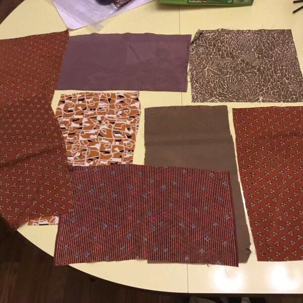 May include: A collection of fabric swatches in different patterns and shades. Features rust-coloured fabric with white floral details, a brown and white geometric pattern, and a striped fabric with blue and red accents. Other swatches are solid colours.