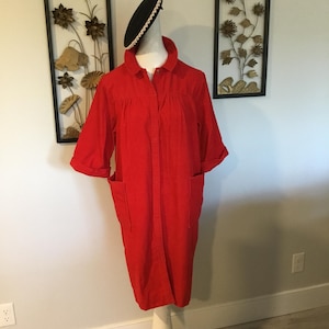 K&ouml;nnte beinhalten: Ein leuchtend rotes, kn&ouml;pfbares Kleid mit kurzen &Auml;rmeln und zwei Vordertaschen. Das Kleid wird auf einer wei&szlig;en Schaufensterpuppe pr&auml;sentiert. Ein schwarzer Hut mit wei&szlig;er Verzierung sitzt auf dem Kopf der Schaufensterpuppe. Wandkunst im Hintergrund.